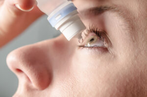 Close up view of young woman applying eye drop to left eye, arti